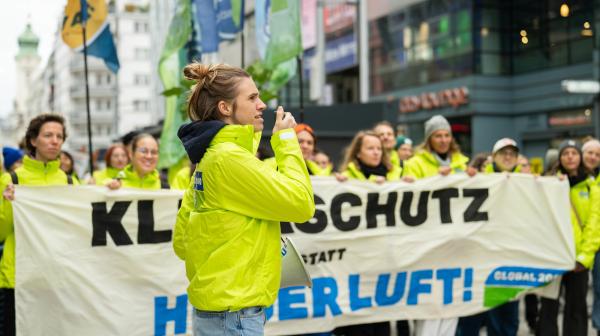 Demonstrierende in GLOBAL 2000 Jacken, im Vordergrund steht eine Person mit Megafon, im Hintergrund sind mehrere Personen die ein Banner halten mit der Aufschrift "Klimaschutz statt heißer Luft"