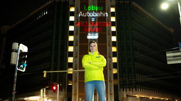 In der Mitte des Bildes steht eine junge Frau mit entschlossenem Blick und verschränkten Armen vor einem Gebäude, dem österreichischen Verkehrsministerium. Die Frau trägt eine Jacke mit einem Global 2000 Logo. Auf das Gebäude hinter ihr ist ein Schriftzug gebeamt "Lobau Autobahn = Milliardengrab!"