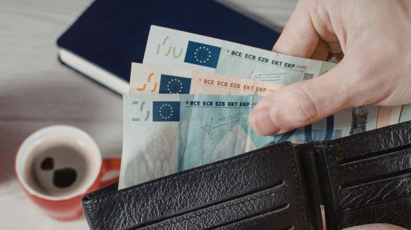 Eine Hand zieht mehrere Euro Geldscheine aus einer Geldbörse, auf dem Tisch dahinter steht ein Kaffee und es liegt ein Notizbuch daneben