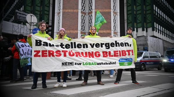 Demonstrierende in Jacken von Global 2000 vor dem österreichischen Verkehrsministerium. Sie halten ein Banner mit der Aufschrift "Baustellen vergehen, Zerstörung bleibt! Nein zur Lobau-Autobahn!"