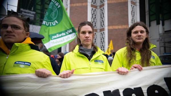 Mehrere Personen in knallgelben Jacken und GLOBAL 2000-Logo mit Banner in den Händen, dahinter Fahnen, auf einer Demo