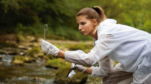 Wissenschaftlerin in weißem Schutzanzug und Handschuhen entnimmt Wasserprobe aus einem Fluss