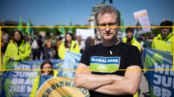 Johannes Wahmüller, Klima- & Energie-Sprecher bei GLOBAL 2000 steht mit verschränkten Armen und entschlossenem Blick vor für Umwelt- und Klimaschutz protestierenden Menschen