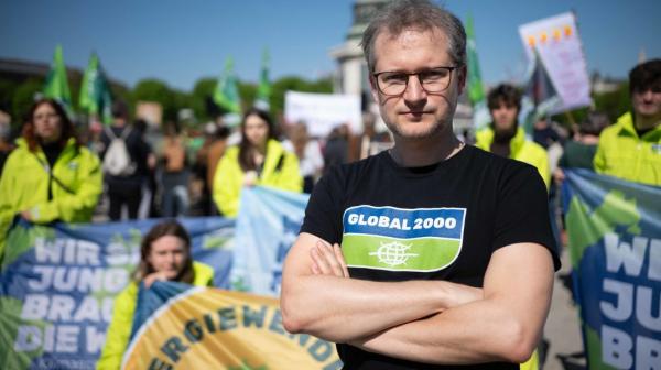 Johannes Wahlmüller von GLOBAL 2000 mit verschränkten Armen vor einer Gruppe von Demonstrierenden in gelben Jacken und mit Fahnen und Bannern.
