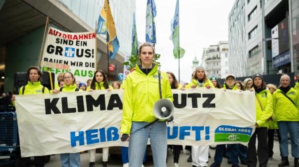 Junge Frau in GLOBAL 2000 Jacke hält Megafon in der Hand, hinter ihr stehen Menschen mit Bannern und Fahnen. Auf einem großen Banner steht "Klimaschutz statt heißer Luft"