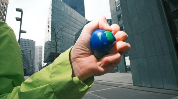 Eine Hand zerquetscht einen Gummiball, der wie eine Erdkugel aussieht. Im Hintergrund sind graue Straßen und Hochhäuser.