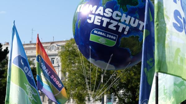 Vor einem Gebäude sind mehrere Fahnen mit dem GLOBAL 2000 Logo, im Zentrum ist eine große aufblasbare Weltkugel mit der Aufschrift "Klimaschutz Jetzt!"