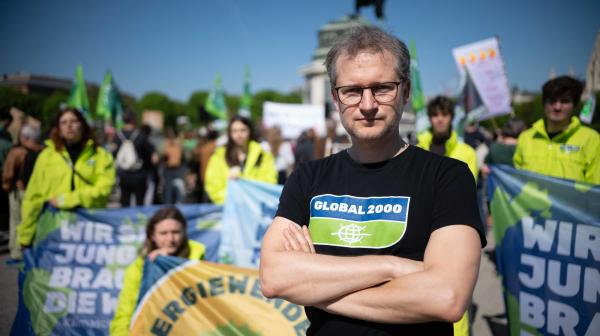 Im Vordergrund steht ein Mann mit verschränkten Armen und einem "GLOBAL 2000" T-Shirt. Im Hintergrund sind Aktivisten mit gelben Jacken und Fahnen zum Thema Klimaschutz und Energiewende