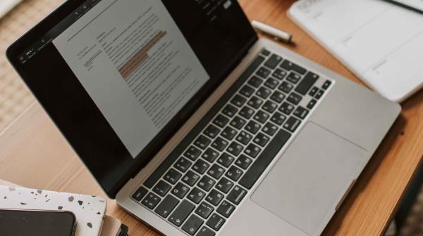 Laptop aufgeklappt auf einem Holztisch mit Block daneben udn STiften