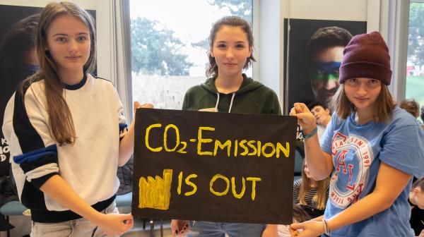 Drei junge Frauen halten ein schwarzes Schild mit gelber Aufschrift 'CO2-EMISSION IS OUT' in einem hellen Raum mit Fenstern