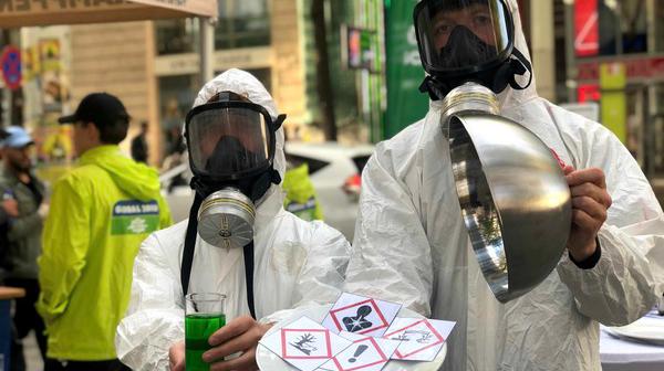 Zwei Personen in weißen Schutzanzügen und Atemschutzmasken halten ein Glas mit grünem Flüssigkeit und ein rundes Metallobjekt, davor ein Papier mit Gefahrensymbolen.
