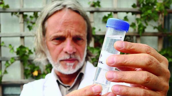 Dr. Helmut Burtscher-Schaden von GLOBAL 2000 im weißen Kittel hält eine durchsichtige Probegefäß mit blauem Deckel vor einem Holzgitter mit Pflanzen
