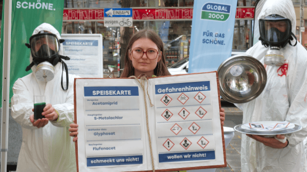 Zwei Personen in Schutzanzügen mit Atemschutzmasken halten links ein grünes Getränk und rechts eine Metallschüssel, dazwischen eine Frau mit Brille hält ein großes Schild mit einer Speisekarte und Gefahrensymbolen, im Hintergrund ein Banner mit der Aufschrift 'GLOBAL 2000 FÜR DAS SCHÖNE'