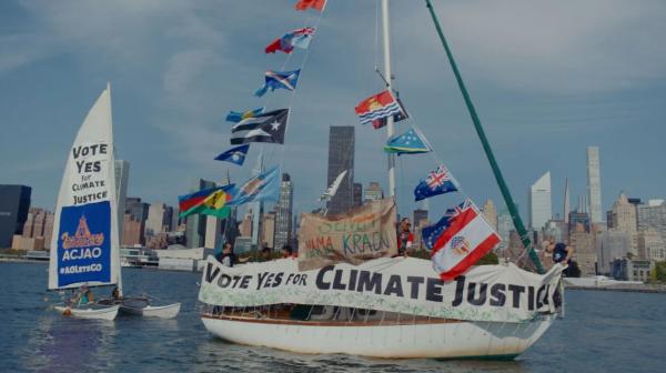 Segelboot mit mehreren Flaggen und Transparenten mit der Aufschrift 'Vote Yes for Climate Justice' vor einer Stadtsilhouette