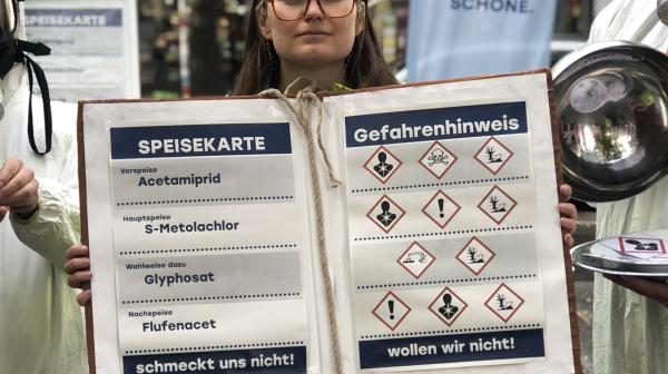 EU-Umweltsprecherin Pitzer hält eine übergroße Speisekarte mit den Namen der giftigen Wirkstoffe und den Gefahrenzeichen