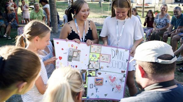 Drei junge Frauen zeigen im Freien ein großes, bunt gestaltetes Plakat mit der Aufschrift 'DANKE SCHÖN' vor einer Gruppe sitzender und stehender Menschen.