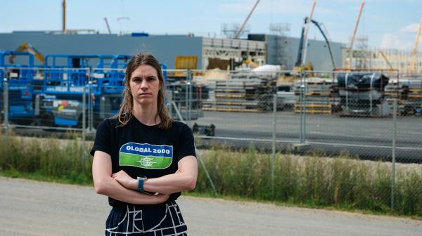 Hannah Keller, mit verschränkten Armen steht vor einem umzäunten Industriegebiet mit Baustellen und Kränen im Hintergrund.